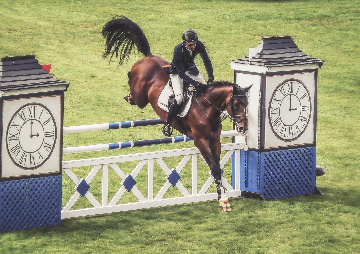 A rider and bay horse clearing a jump between clock-tower standards